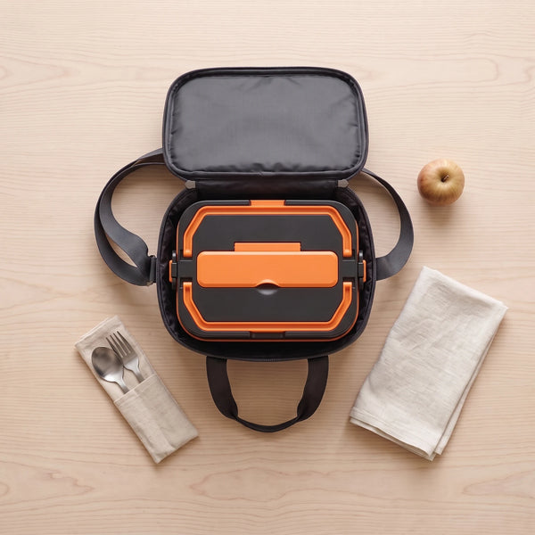 A top-down flat lay of a black insulated bag open on a pale wooden table with the closed black-and-orange lunch box packed inside, a cloth roll with a steel fork and spoon, a folded napkin and a small apple beside it