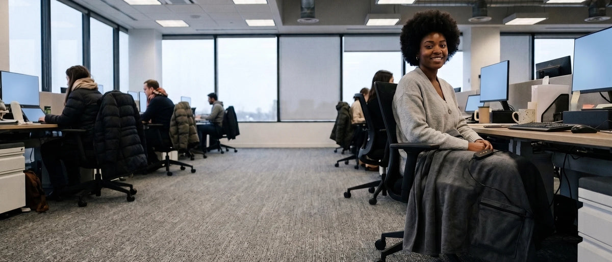 An open-plan office on a grey winter morning with colleagues in puffy jackets out of focus on the left, and a smiling young woman in a light-grey cardigan at a desk on the right with a graphite-grey blanket over the lap and legs and a mug beside the keyboard