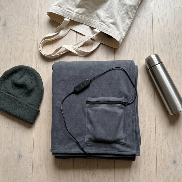 A top-down flat lay on a pale wooden floor of a graphite-grey blanket folded into a thick rectangle with a zippered pocket corner on top, a coiled black cable and a small black controller beside it, a canvas tote, a knit beanie and a steel flask
