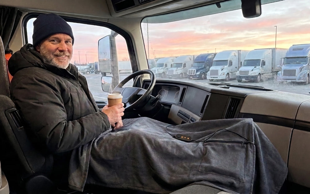 The driver seat of a parked truck cab at dawn with a smiling man in a navy beanie, a graphite-grey blanket over the legs and a steaming paper cup in the hand, a row of parked trucks and a pink-orange sky through the windshield